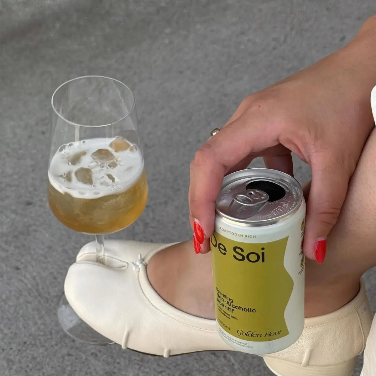 Hand holding De Soi alcohol-free beverage can next to a glass of golden drink with ice