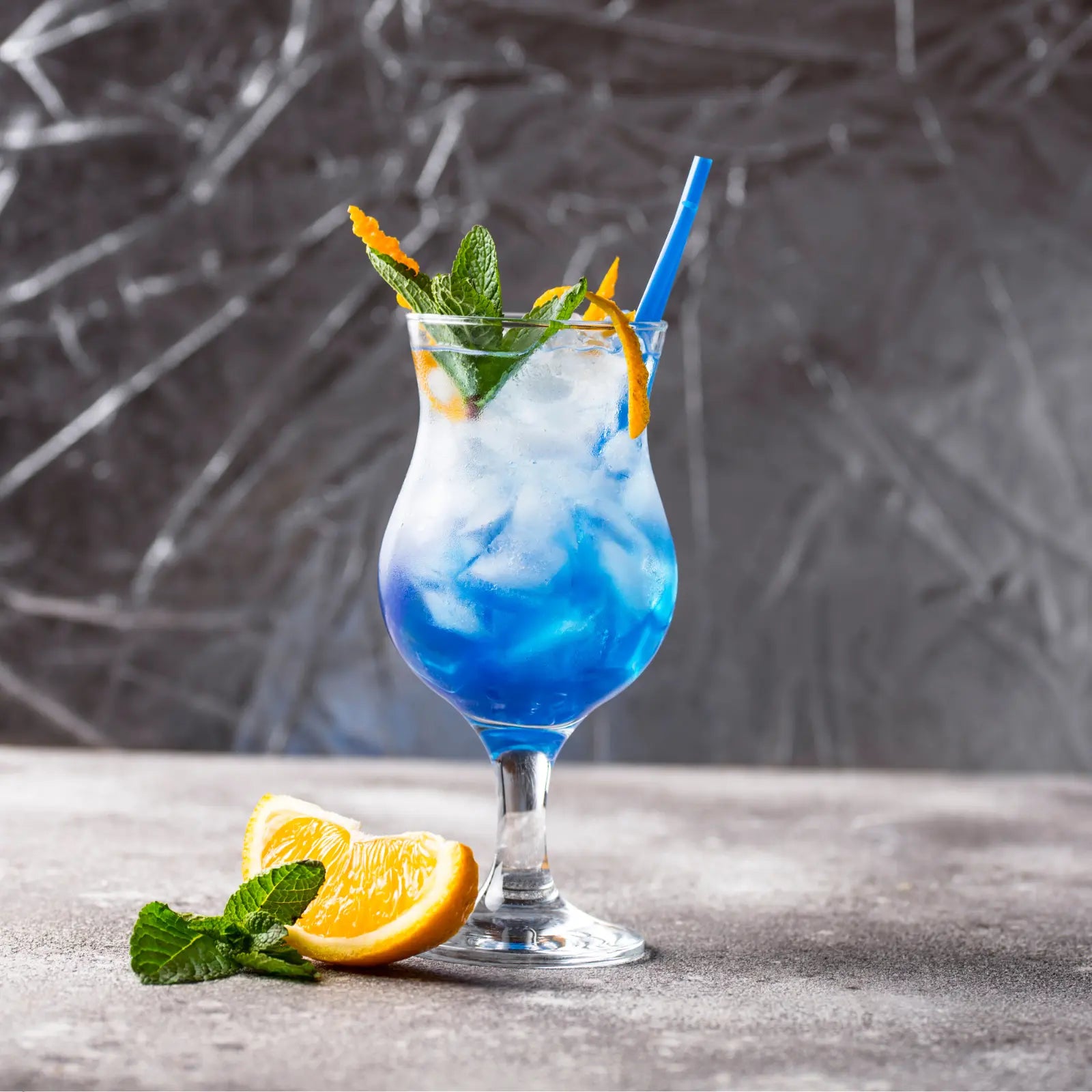 Blue lagoon mocktail garnished with orange peel, mint, and a straw in a curvy glass on a gray textured surface