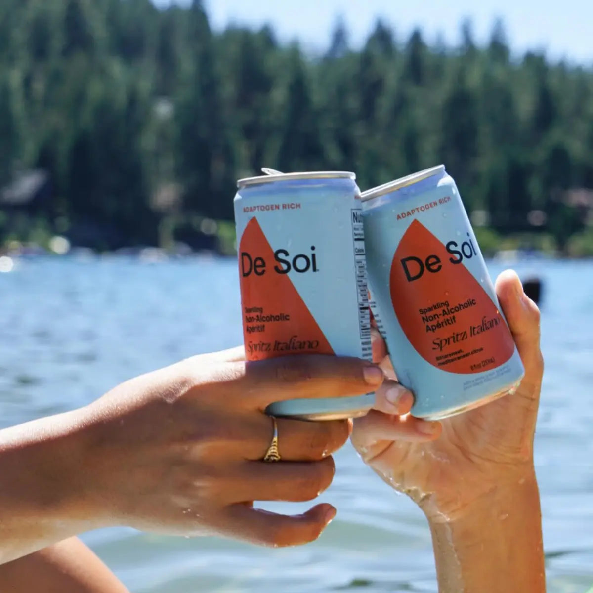 Two hands toasting De Soi Spritz Italiano non-alcoholic aperitif cans by a forest lake.