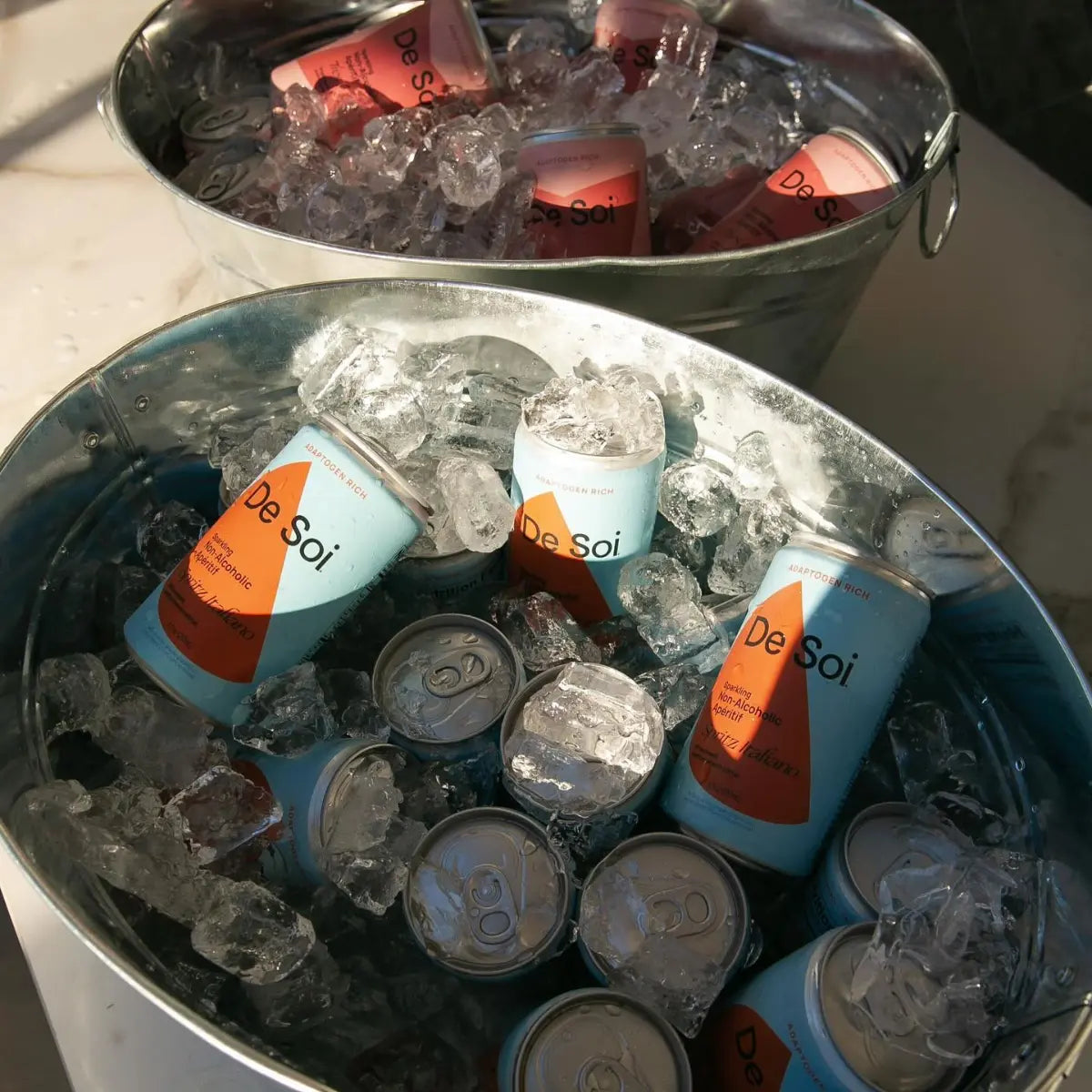 Cans of De Soi non-alcoholic aperitif chilling on ice in metal party buckets.
