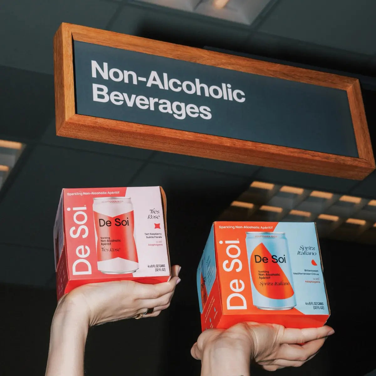 Hands holding De Soi non-alcoholic cocktail packs beneath a wooden "Non-Alcoholic Beverages" sign