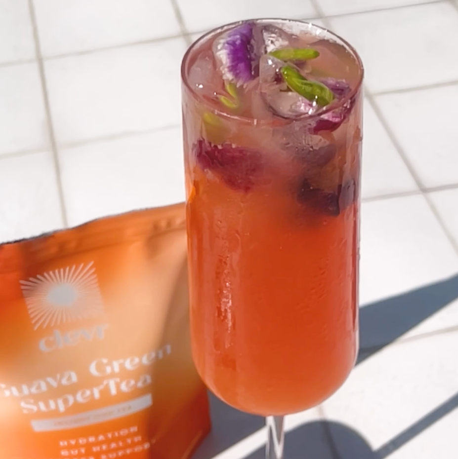 Non alcoholic rosé spritz with edible flowers beside a Clevr Guava Green pouch on sunlit tile.