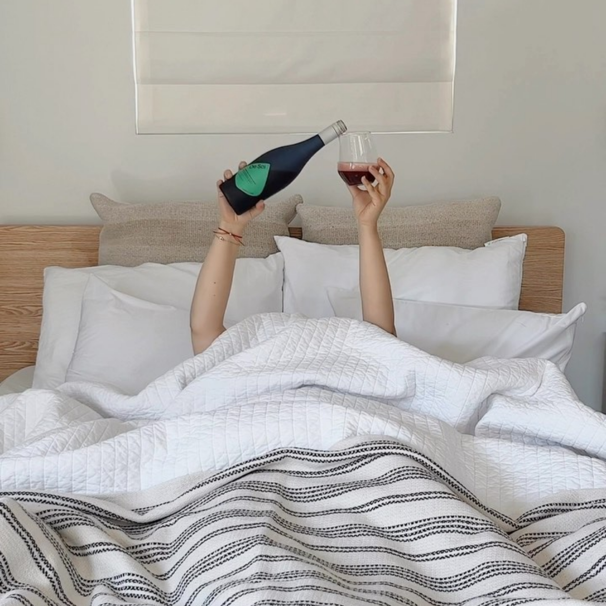 Person lying in bed under white blankets, holding a bottle of non-alcoholic aperitif and a glass above the covers