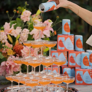 A pyramid of cocktail glasses being filled with orange De Soi non-alcoholic drinks, surrounded by floral decorations and canned drinks