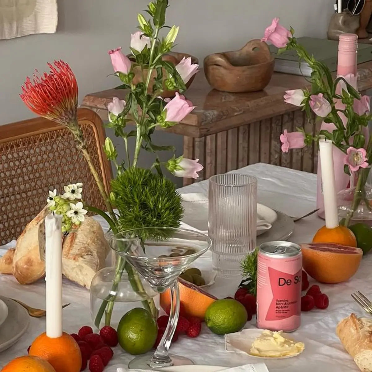 A table set with a De Soi non-alcoholic drinks, fresh fruit, bread, flowers, and candles for a vibrant dining display