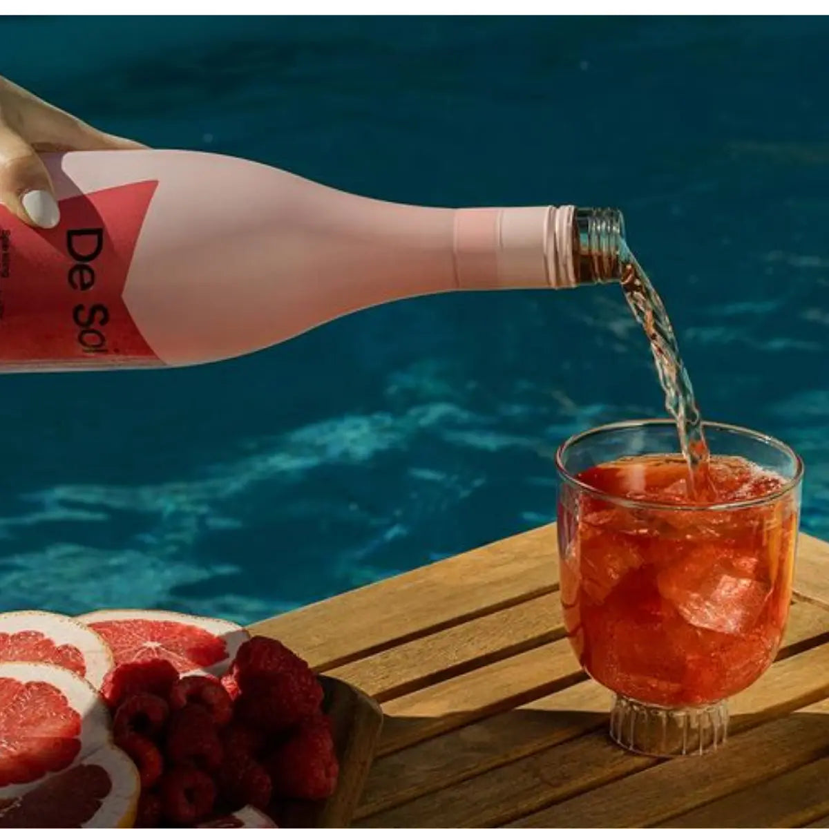 Pouring a De Soi botanical non-alcoholic drink into a glass with ice, set beside fresh raspberries and grapefruit by a pool.