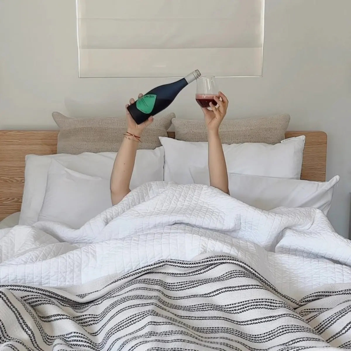 Hands emerging from bed covers pour a De Soi non-alcoholic aperitif into a glass.