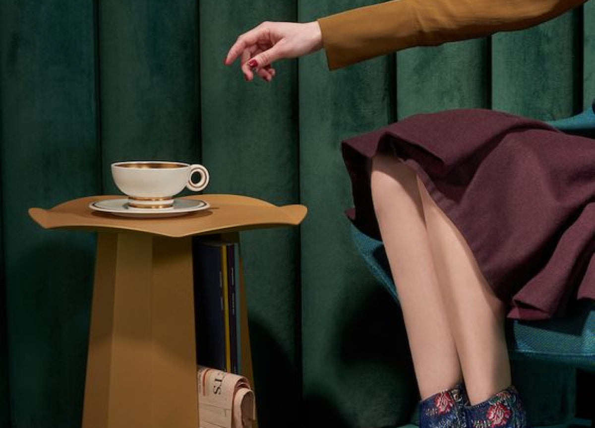 Hand in mustard sleeve reaching toward a gold-rimmed teacup on a side table.
