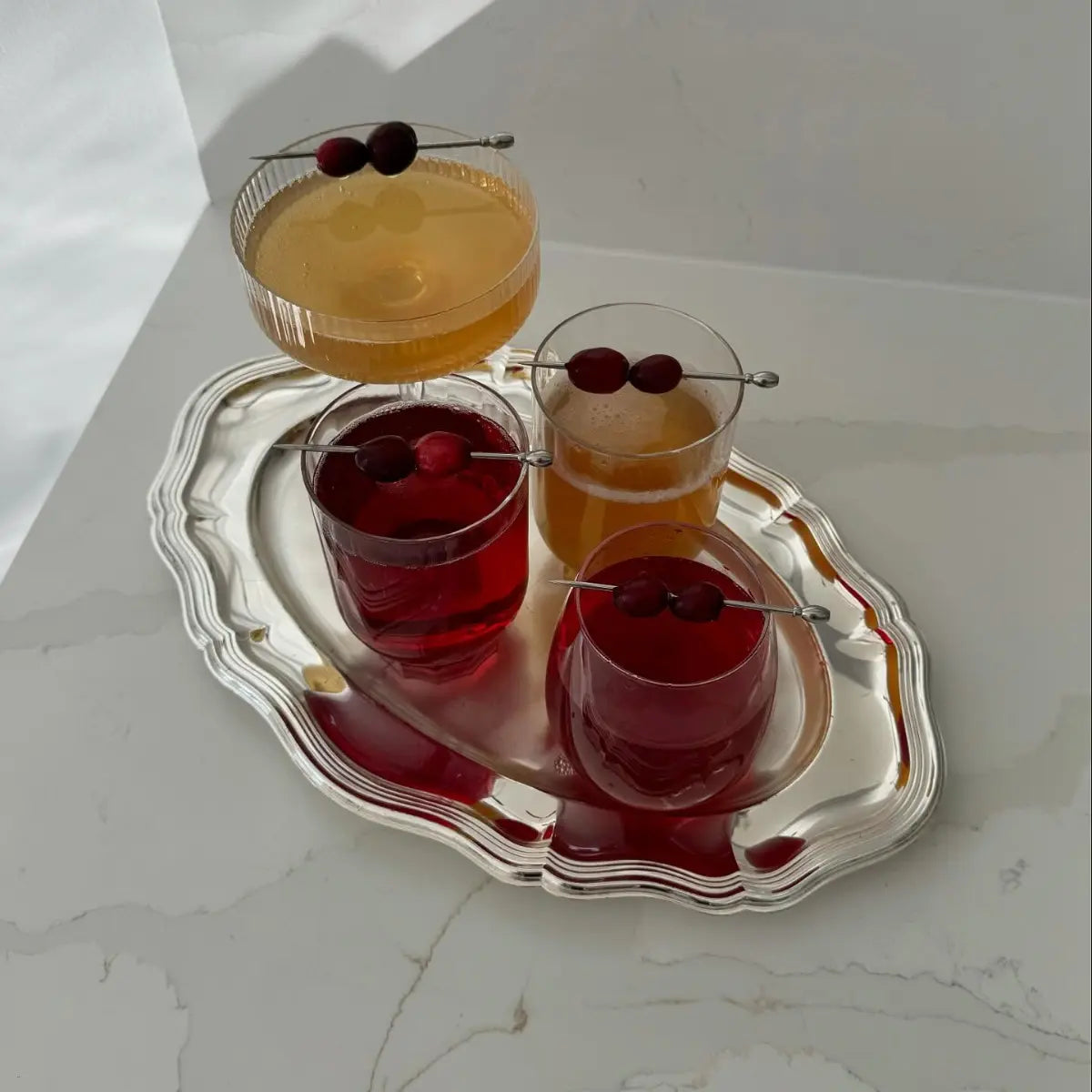 Non-alcoholic drinks garnished with cranberries arranged on a silver tray with natural light on a marble surface.