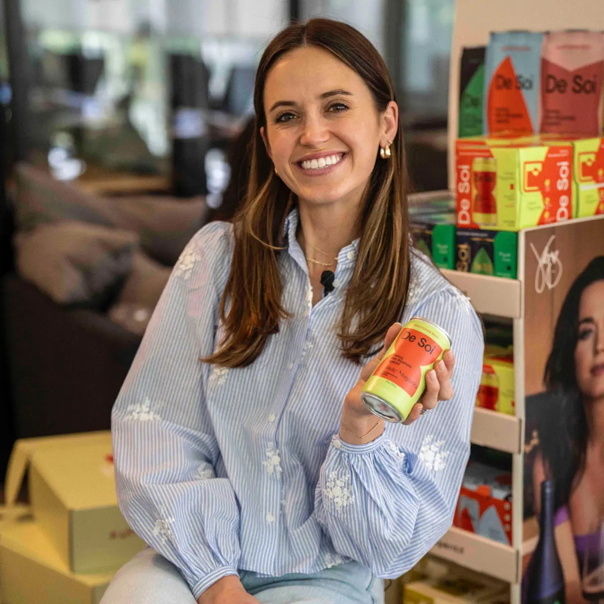 Non-alcoholic aperitif brand ambassador smiling while holding a De Soi can in front of product display.