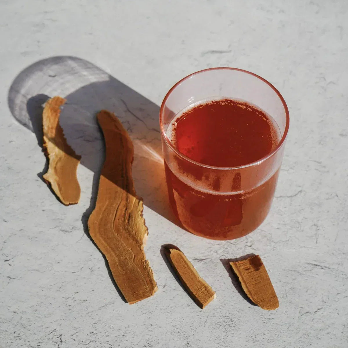 Reddish adaptogen mocktail in a glass beside dried root pieces on a sunlit surface.