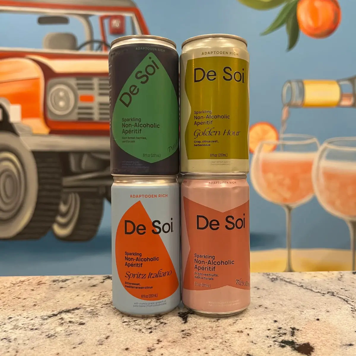 Modern apéritifs in colorful De Soi cans stacked in front of a mural with a vintage car and cocktails