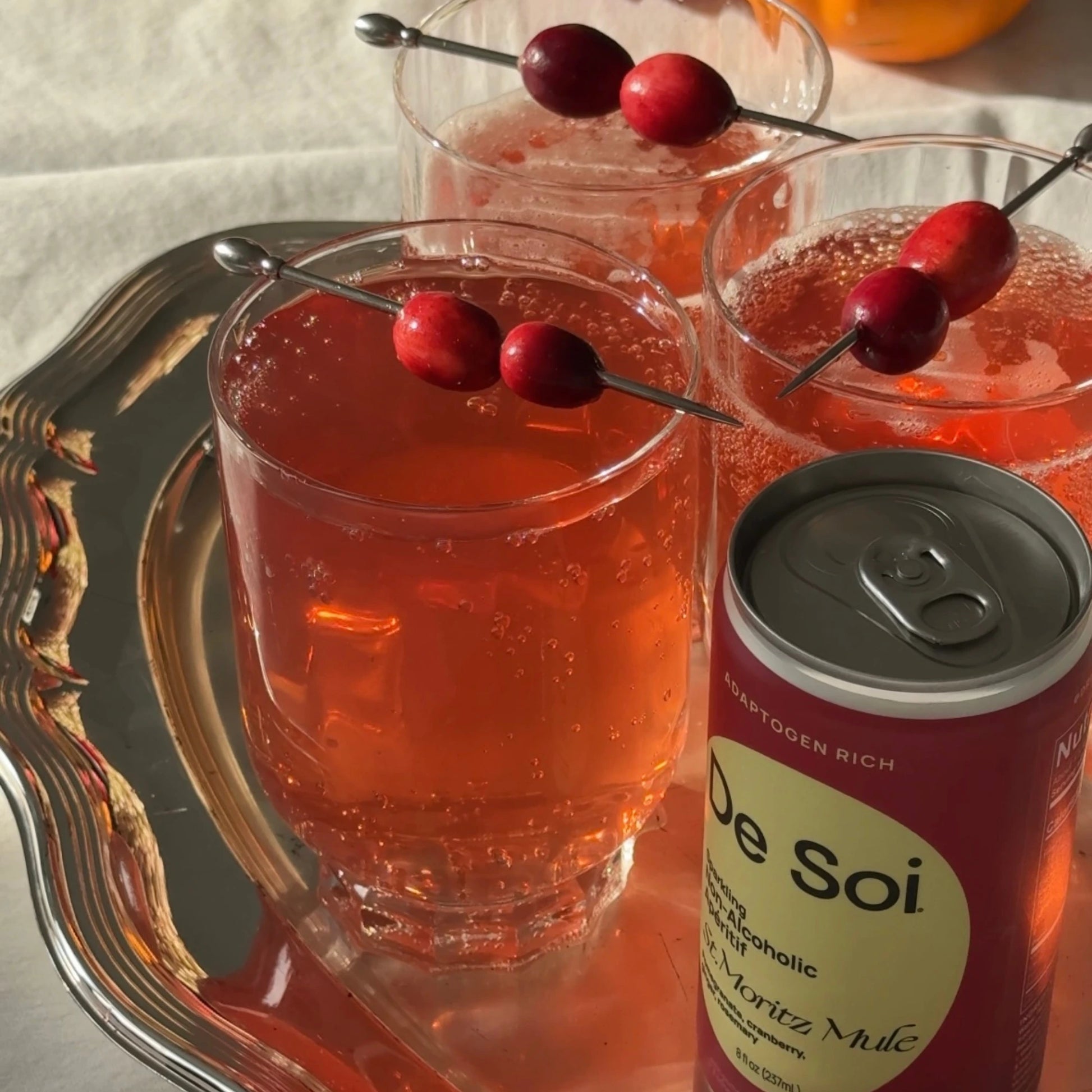 Two glasses of sparkling red cocktail with cranberry garnish on a silver tray alongside a De Soi St. Moritz Mule can.