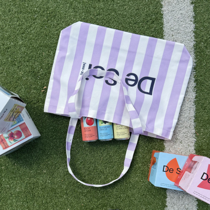 Lilac striped De Soi canvas tote bag holding drink cans on grass.