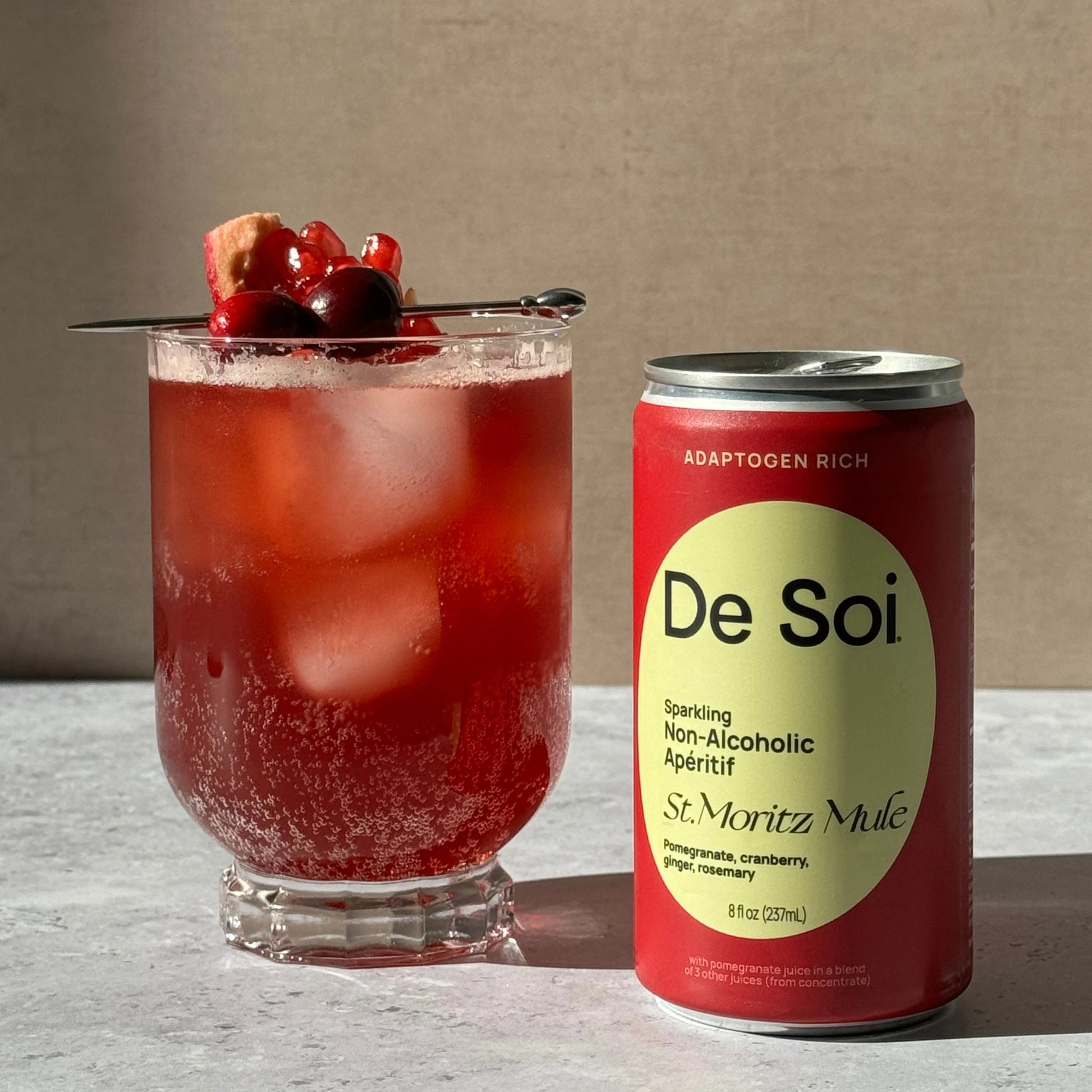 De Soi St. Moritz Mule can beside a tall glass of sparkling red cocktail with ice and pomegranate cherry garnish on a light stone surface.