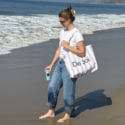 Person on a sandy beach carrying the lilac striped De Soi canvas tote over one shoulder.