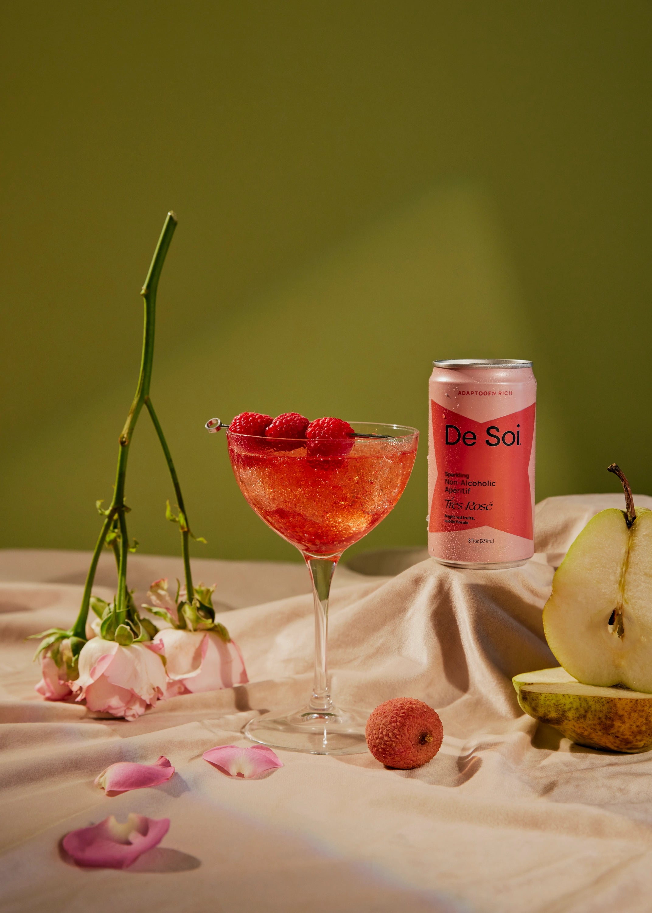De Soi Très Rosé non-alcoholic sparkling aperitif in a pink can beside a coupe glass topped with fresh raspberries, styled with pear and rose petals.