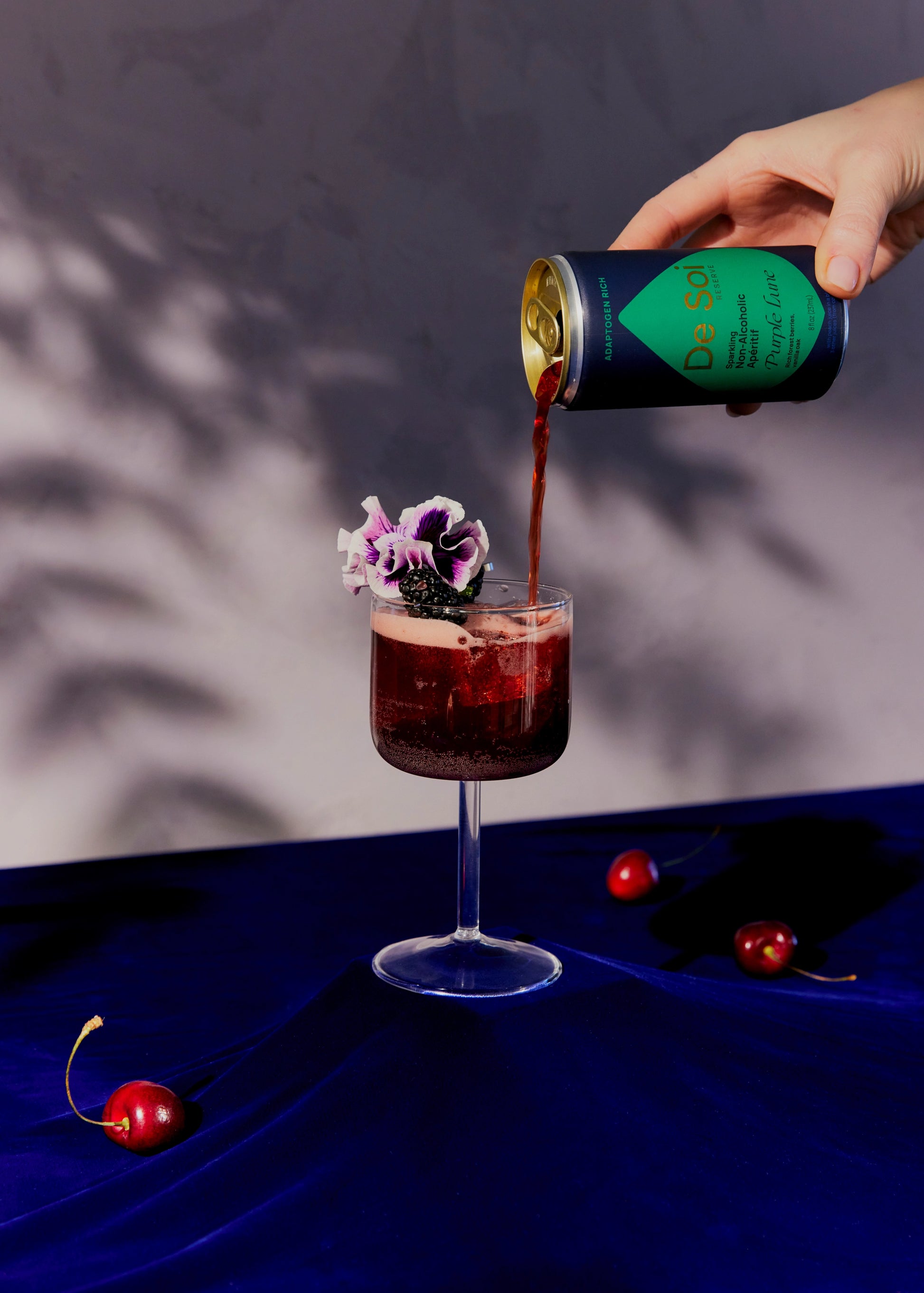 De Soi Purple Lune non-alcoholic aperitif poured from the can into a coupe glass garnished with purple flowers and fresh cherries.