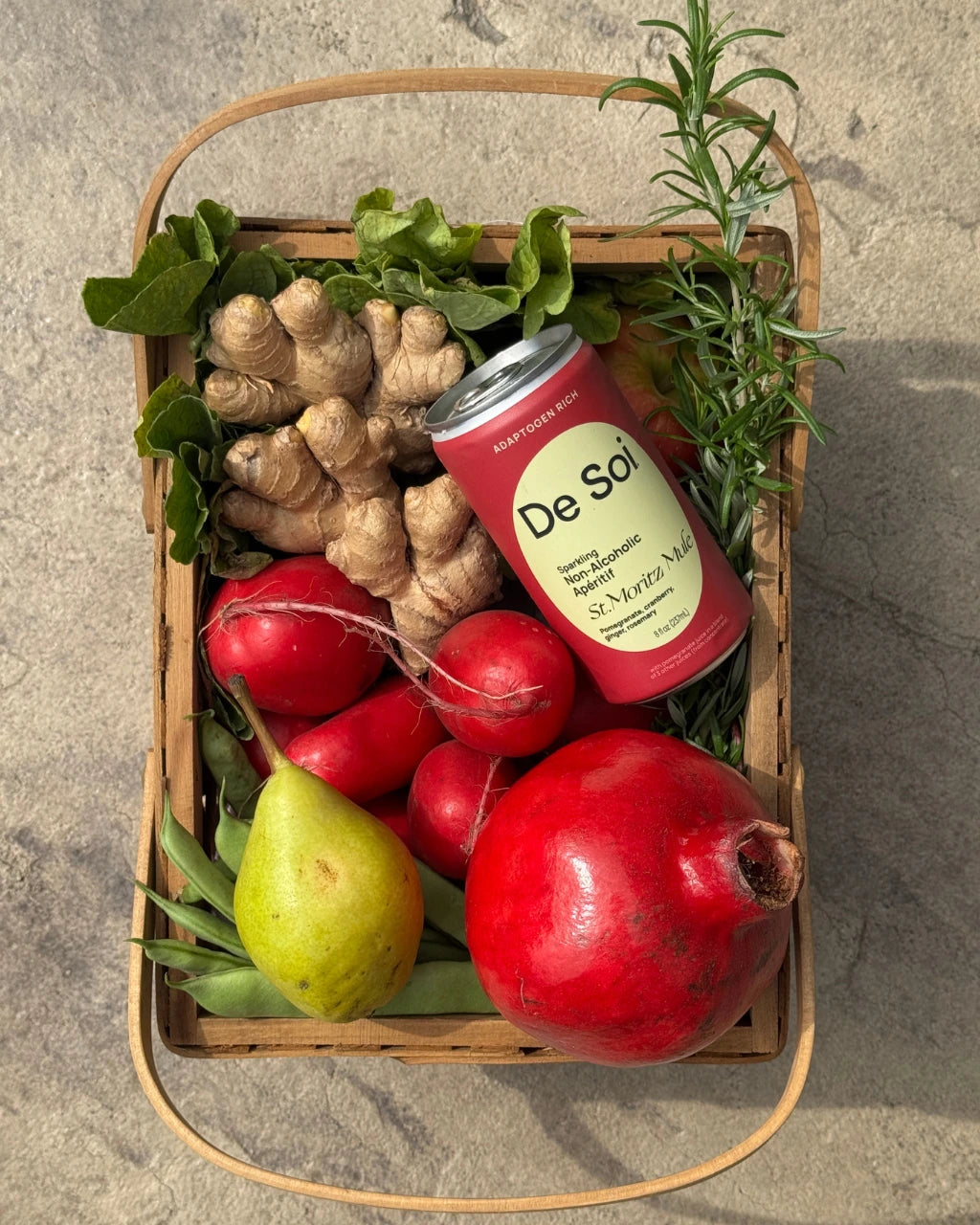 De Soi St. Moritz Mule can nestled in a wicker basket with fresh ginger root, pomegranates, pear, and rosemary on a stone surface.