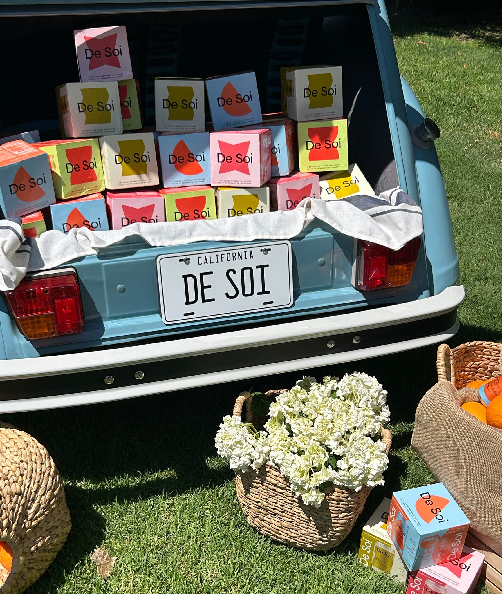 Open car trunk with De Soi 4-packs, a license plate, and a basket of flowers on grass.