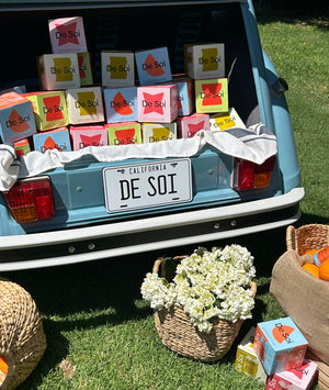 Open car trunk with De Soi 4-packs, a license plate, and a basket of flowers on grass.