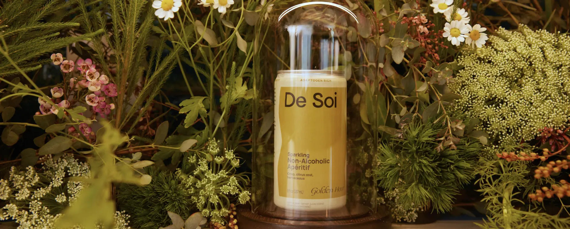De Soi Golden Hour non-alcoholic aperitif can under glass cloche amid wildflowers and greenery.